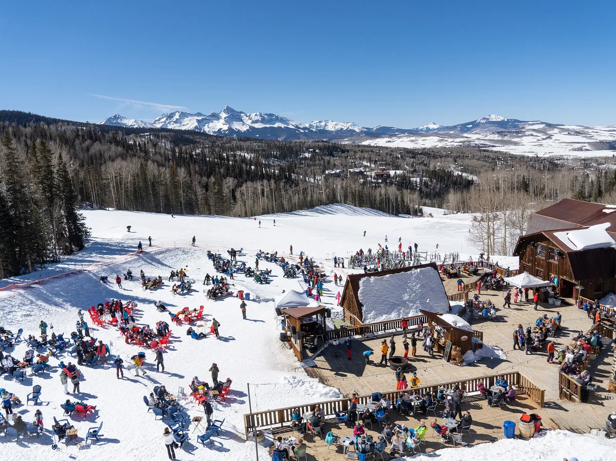 A lively scene of people skiing on snowy slopes with wooden buildings and outdoor seating areas visible, surrounded by mountain vistas. Groups are seen relaxing and socializing, enjoying both sport and après-ski moments in the sun.