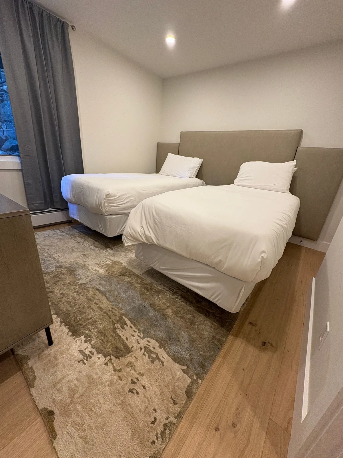 The photo features two neatly arranged twin beds with soft bedding and a decorative rug between them. Grey blackout curtains hang at the window, and the overall look is tranquil and comfortable—ideal for families or friends. The space exudes flexibility and thoughtful touches for a restful night.