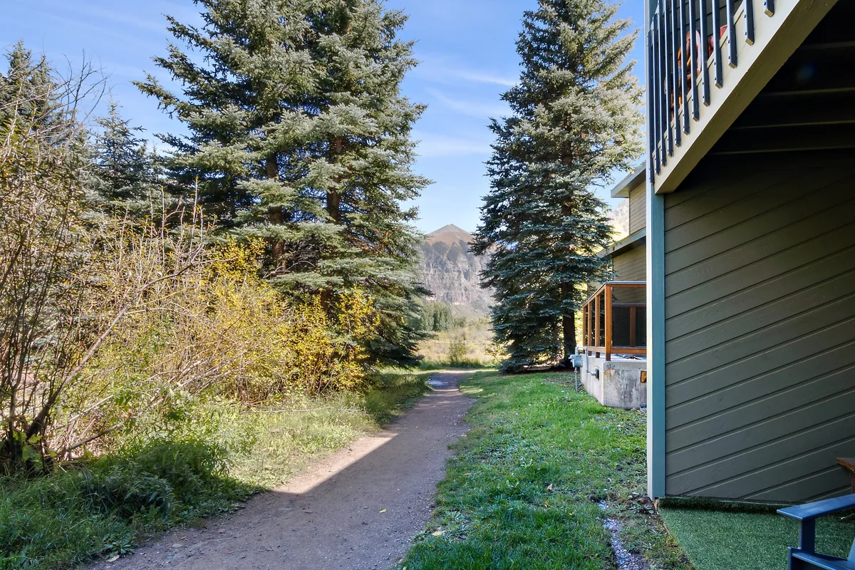 A scenic exterior view featuring a pathway surrounded by lush greenery, with a mountain backdrop and a wooden deck area visible. The image suggests immediate access to trails and nature, with a serene and inviting atmosphere for outdoor activities such as walking, hiking, or relaxing outdoors.