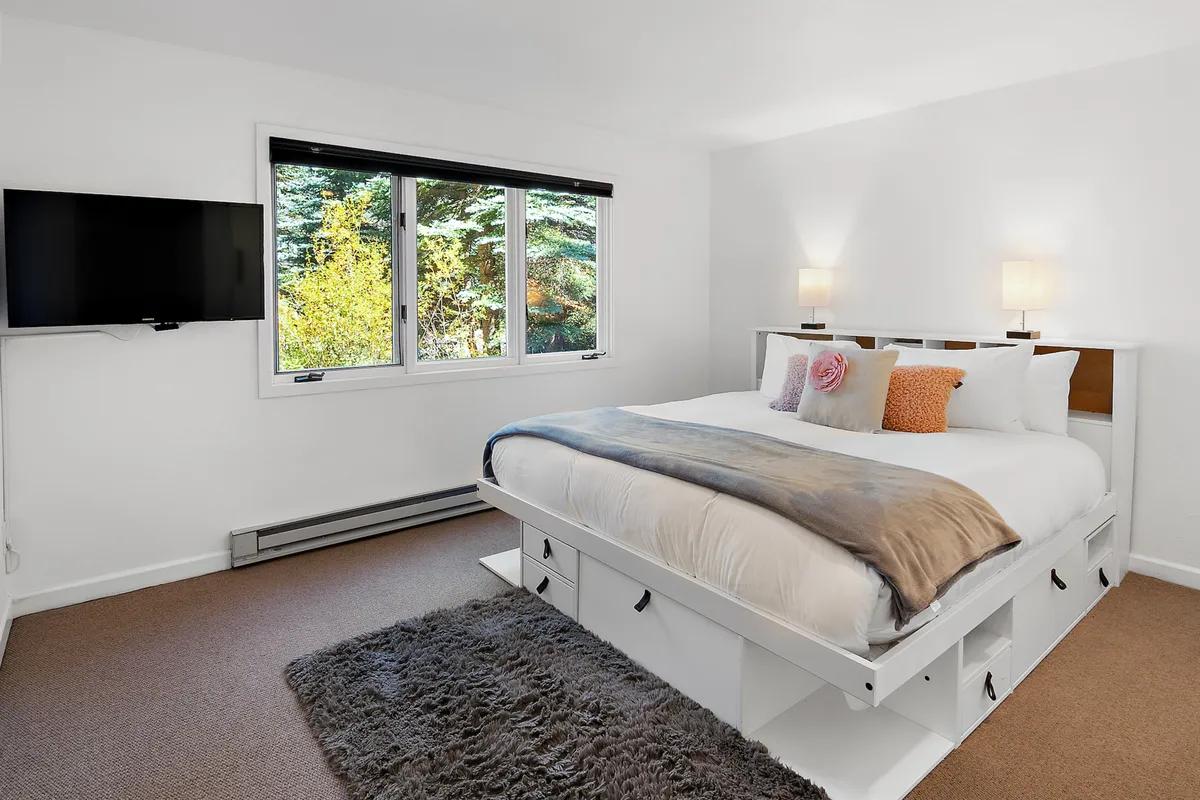 This image displays a large, beautifully made bed with decorative pillows set against a wall, a large window framing a serene nature view, wall-mounted television, and elegant wall sconces. The bedding appears plush and inviting, and natural light bathes the room, highlighting the tranquil atmosphere.