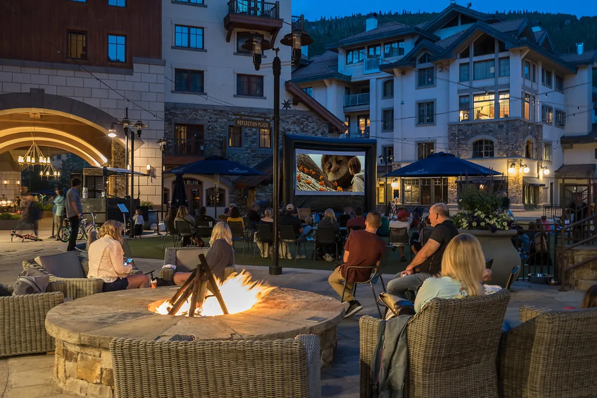 A cozy outdoor gathering around a fire pit, with people watching a movie projected on an outdoor screen under the night sky. The group is surrounded by comfortable seating and soft lighting, creating a festive and communal vibe.