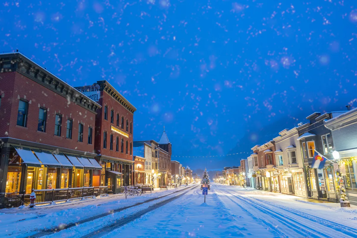 A snow-covered street lined with festively decorated buildings and a Christmas tree in the distance. The mood is warm and inviting, capturing the community spirit during seasonal events and festivals.