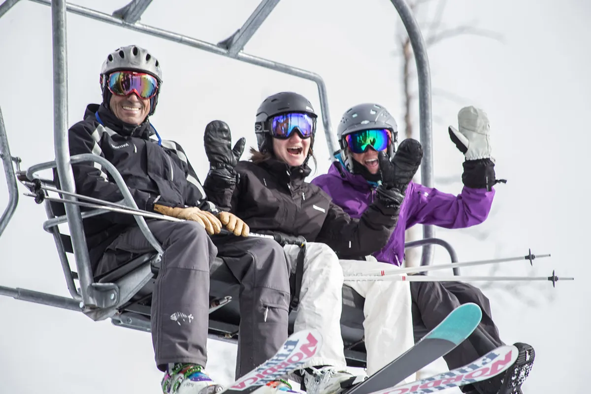 Three skiers ride a chairlift above a snow-covered landscape, dressed in bright ski gear. The scene is joyful, emphasizing shared experiences and the anticipation of an exciting day on the slopes.