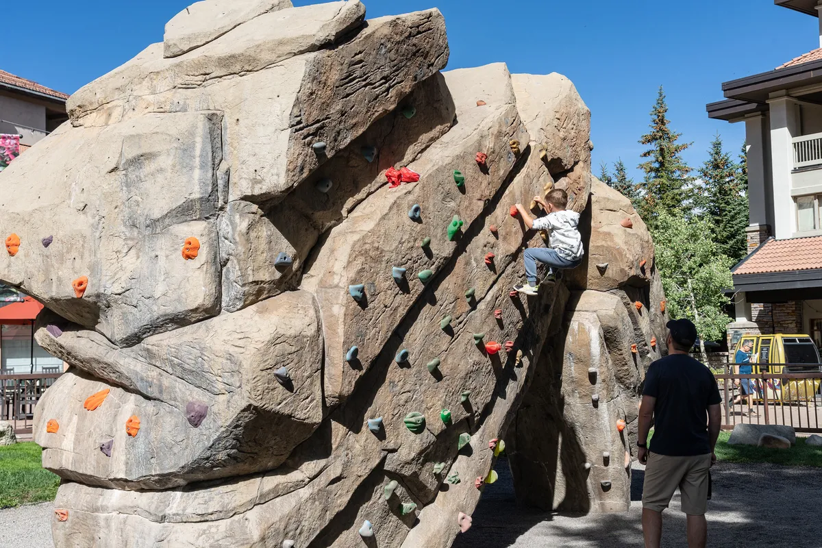 A close-up exterior shot featuring a young child climbing on a public climbing wall with the guidance of an adult. The background includes green space, suggesting a family-friendly park environment ideal for outdoor play and bonding.