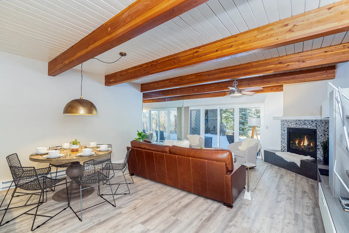 A cozy interior shot focusing on the dining table set for a meal, with a leather sofa and a glowing fireplace in the background. The ambiance is warm and perfect for gatherings, hinting at family dinners, game nights, or casual relaxation by the fire.