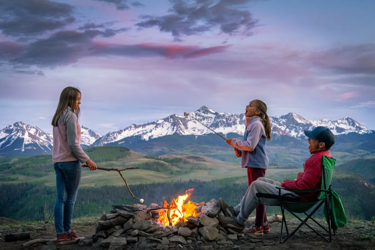 A lively scene showing children roasting marshmallows with a father figure around a campfire, set against a backdrop of snow-capped mountains. The group is clearly enjoying each other's company outdoors, surrounded by natural beauty.