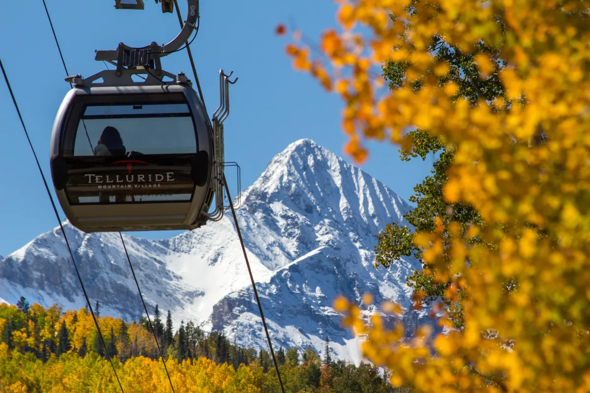 A gondola glides above vibrant autumn foliage with a snow-capped mountain in the background. The scene captures the thrill of ski season, the beauty of changing seasons, and the convenience of being near gondola access for both winter and summer adventures.