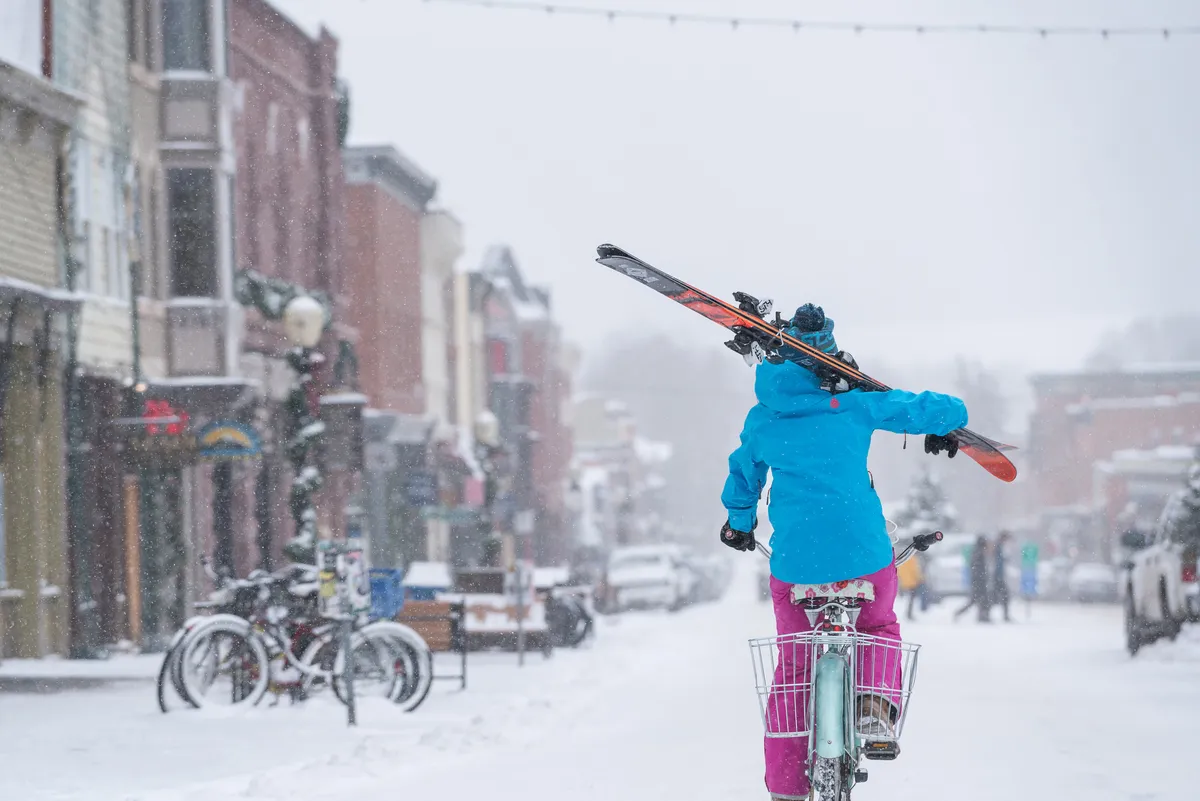 A person rides a bicycle with skis strapped on, navigating a snow-covered street lined with colorful buildings. The scene combines action and local flair, with the festive energy of people exploring town, lively facades, and hints of adventure.