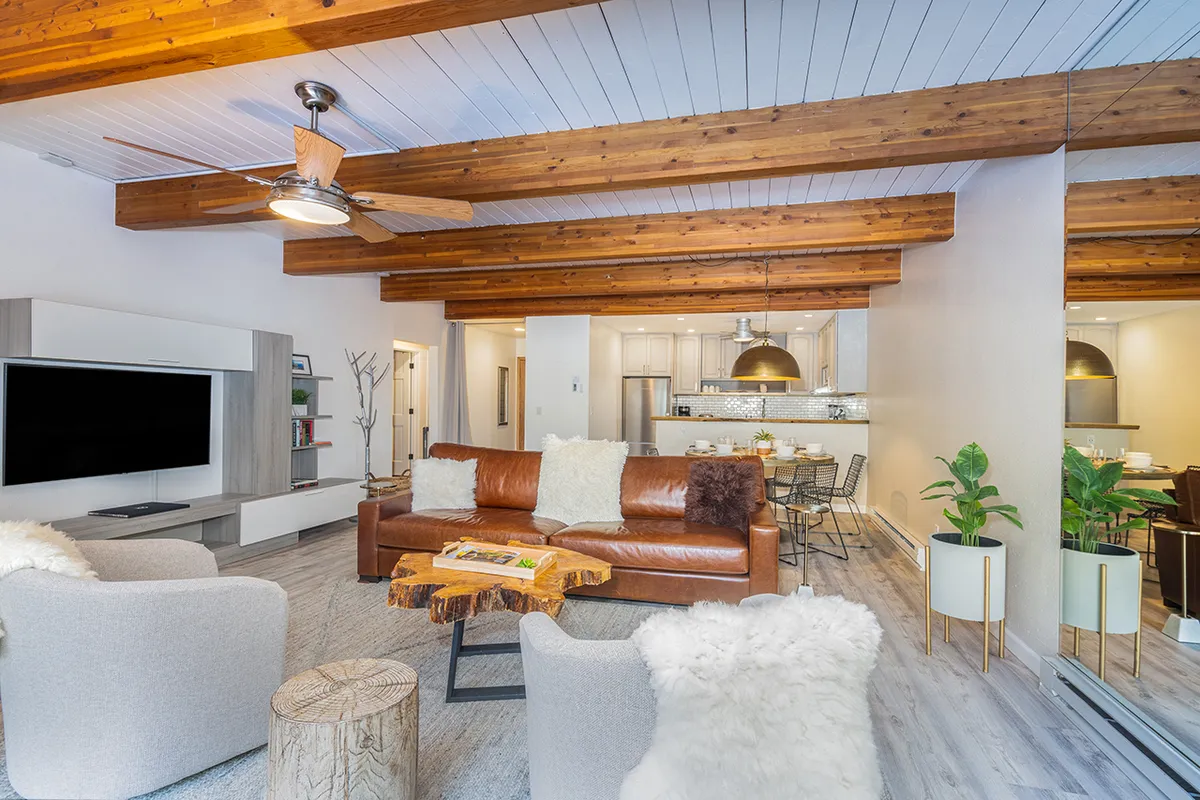 This image features a warm, inviting open-concept space with a leather sofa, coffee table with magazines, wooden beams on the ceiling, green plants adding a homey touch, and a modern kitchen visible in the background. It perfectly captures the social heart of the condo, highlighting both the living area and kitchen in one harmonious view.