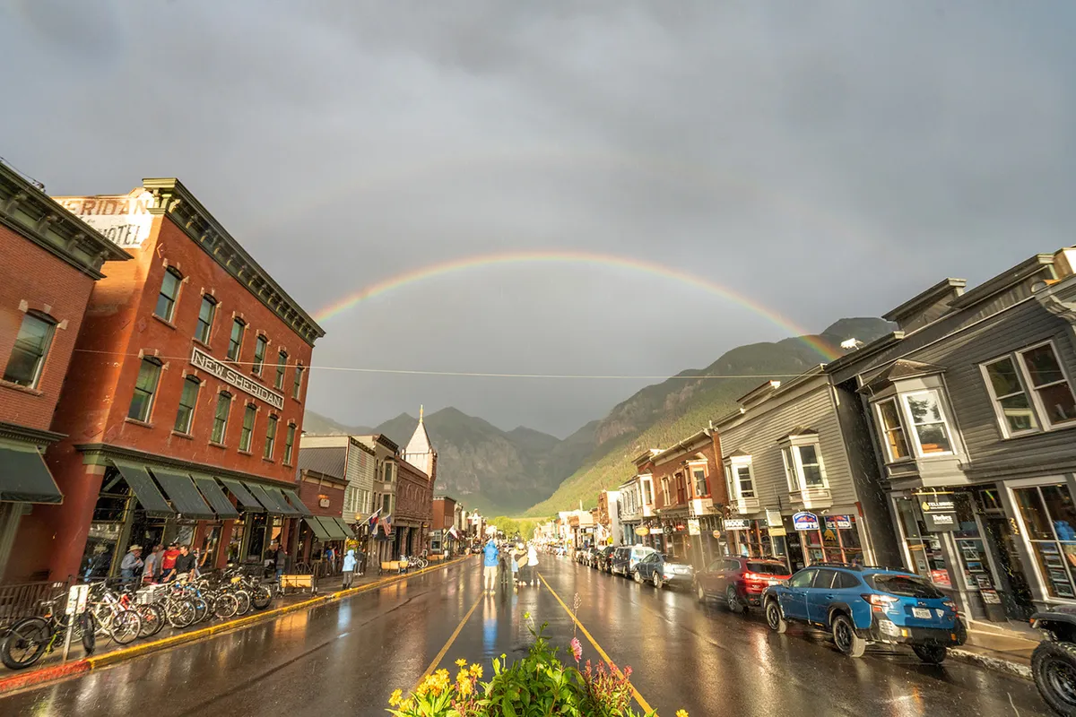 This image shows Telluride's charming historic buildings lining a wet pavement, with a vibrant rainbow arching overhead and the silhouette of distant mountains in the background. The scene is lively yet tranquil, capturing a quintessential moment of small-town exploration and Main Street ambiance.
